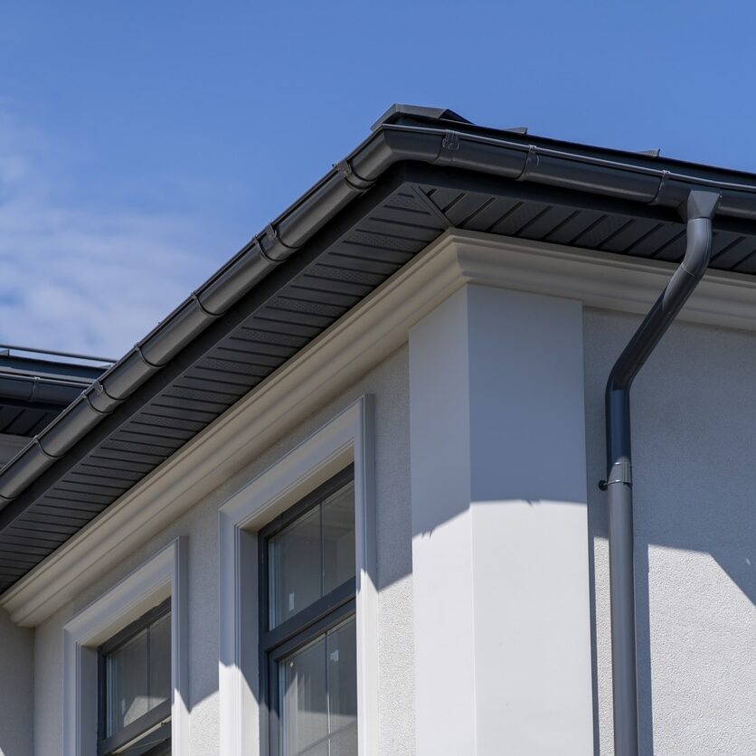 Corner of house with windows, new gray metal tile roof and rain gutter. Metallic Guttering System, Guttering and Drainage Pipe Exterior